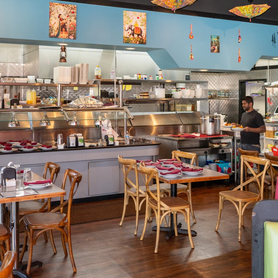 Colorful restaurant dining room with teal walls, Indian artwork, set tables, open kitchen, and a chef carrying plates.