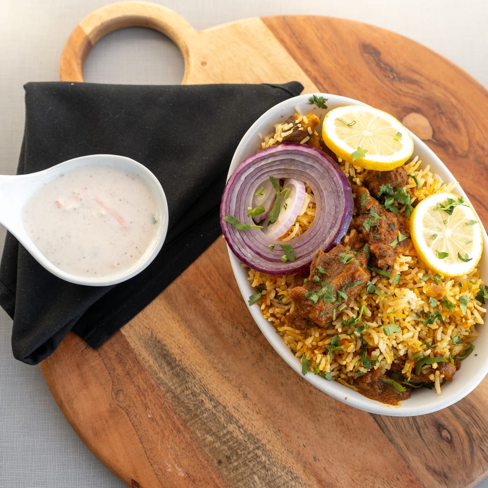 Spiced biryani with meat, red onion, lemon slices, and cilantro served beside creamy raita on a wooden board.