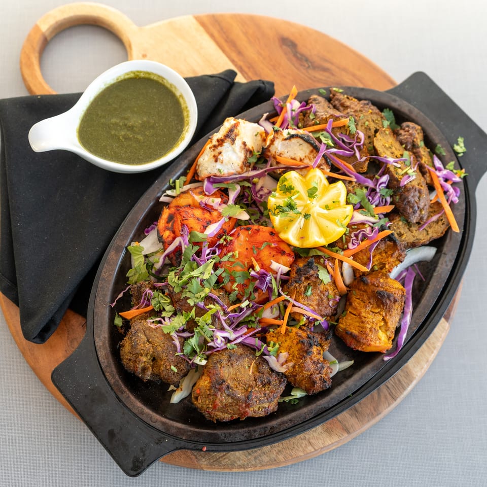 Grilled mixed-meat platter garnished with cabbage, carrots, cilantro, and lemon, served with green chutney on a wooden board.