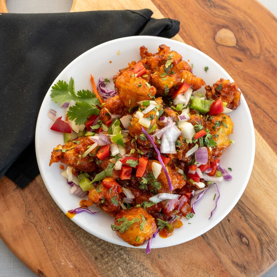 Plate of crispy sauced fritters topped with chopped onions, peppers, cabbage, and fresh cilantro on a wooden table.