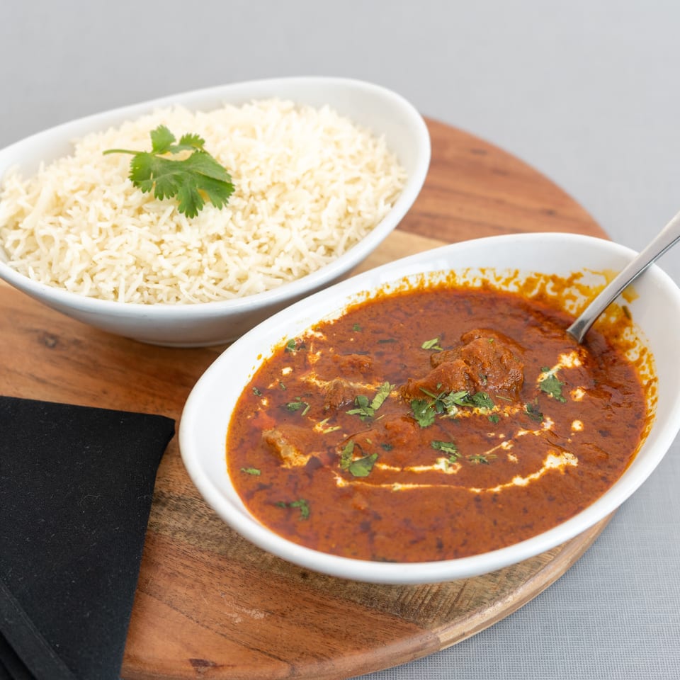 White basmati rice garnished with cilantro sits beside creamy red curry in serving bowls on a wooden board.