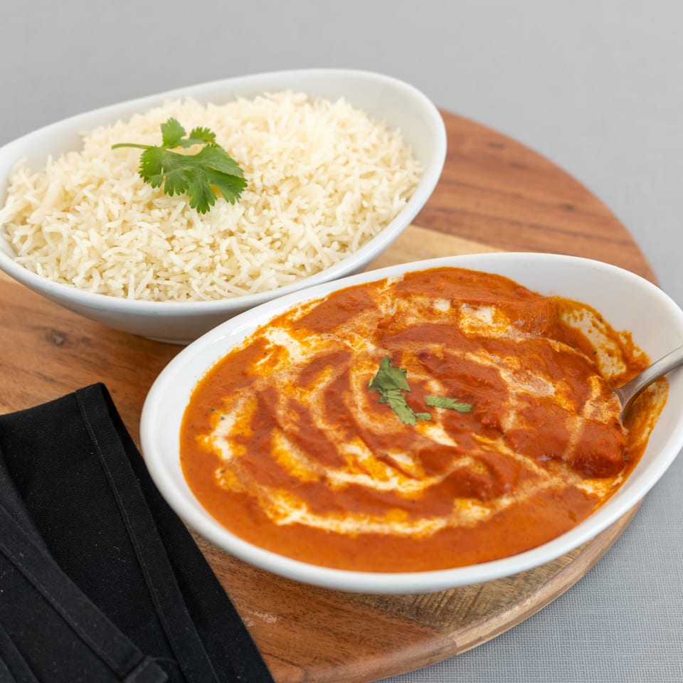 White basmati rice garnished with cilantro beside creamy red curry in serving bowls on a wooden board.