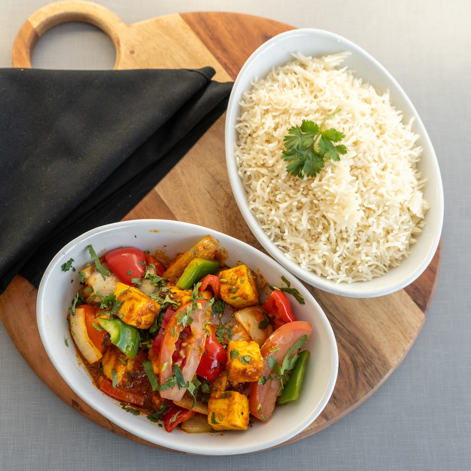Overhead view of spiced paneer with peppers and tomatoes beside basmati rice, garnished with cilantro on a wooden board.