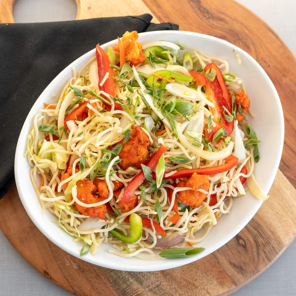 Bowl of noodles with crispy orange chicken, shredded vegetables, bell peppers, cabbage, and scallions on a wooden serving board.