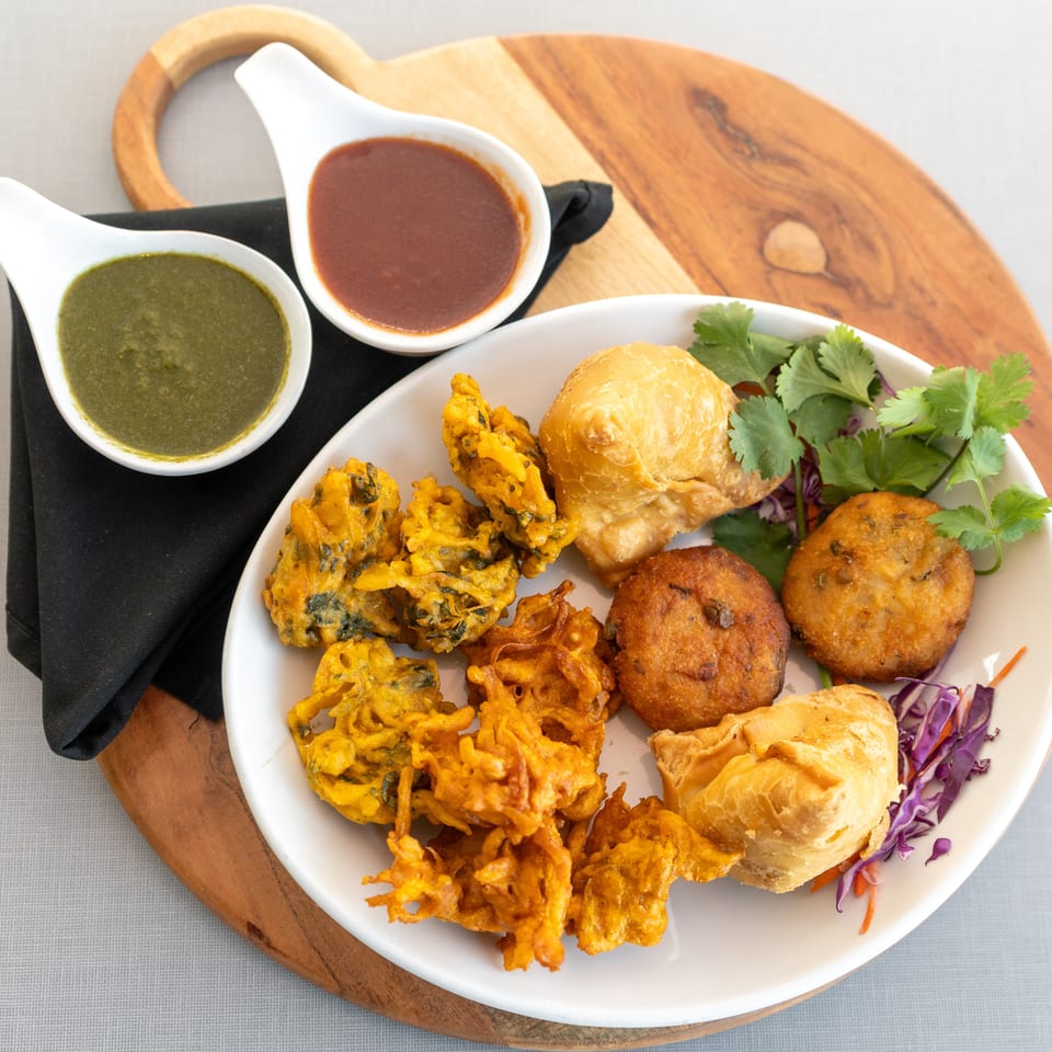 Indian appetizer platter with pakoras, savory pastries, chutneys, cilantro, and shredded cabbage on a wooden serving board.
