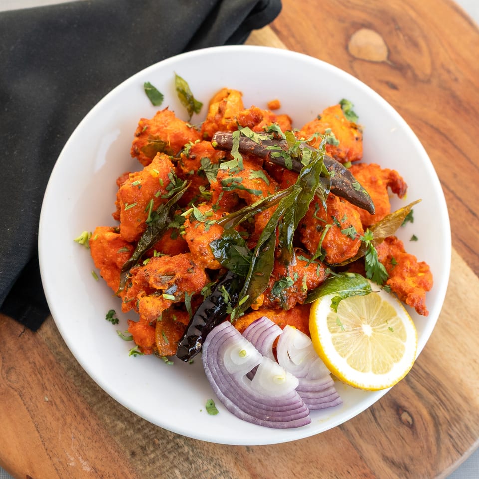 Plate of crispy red-spiced fritters garnished with curry leaves, cilantro, lemon, and sliced red onion on a wooden table.