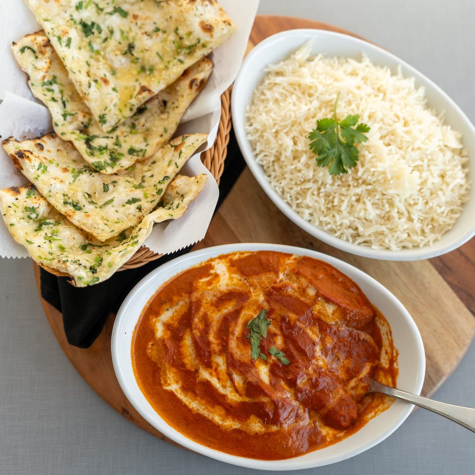 Indian meal featuring garlic naan, basmati rice garnished with cilantro, and creamy red curry served on a wooden board.