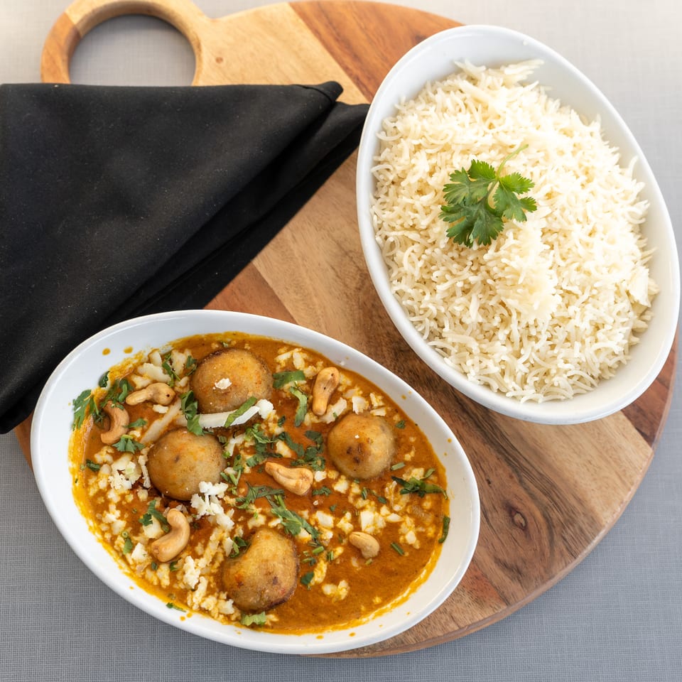 Overhead view of creamy curry with vegetable dumplings, cashews, and cilantro beside basmati rice on a wooden serving board.