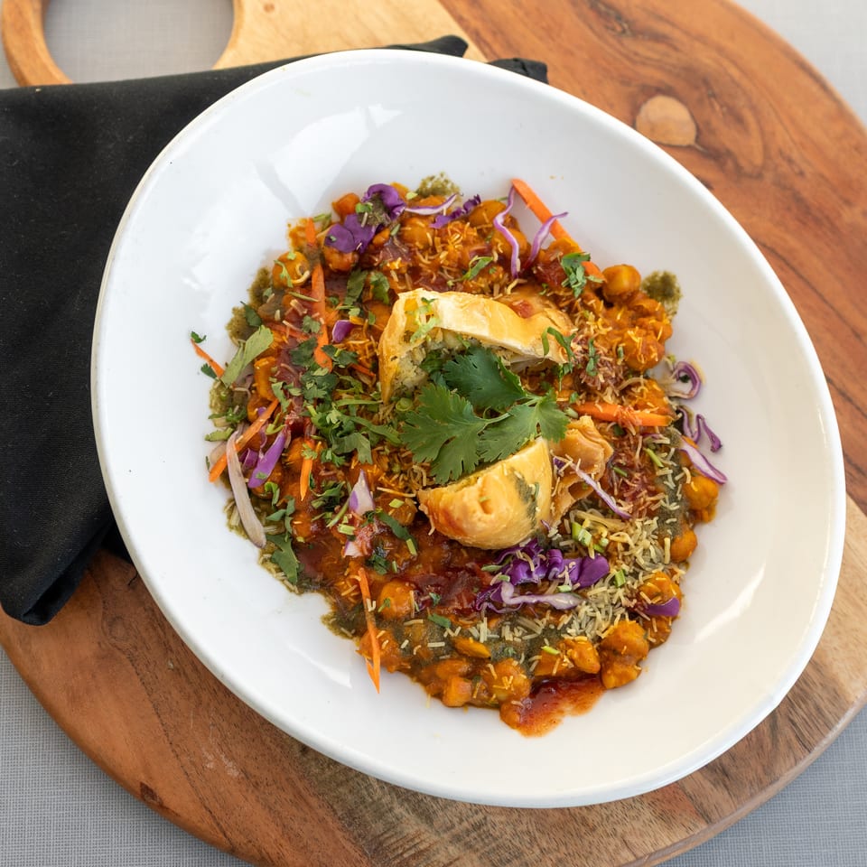 Colorful Indian chaat platter topped with crispy pastry, sev, cilantro, shredded cabbage, carrots, and chutneys on a white plate.
