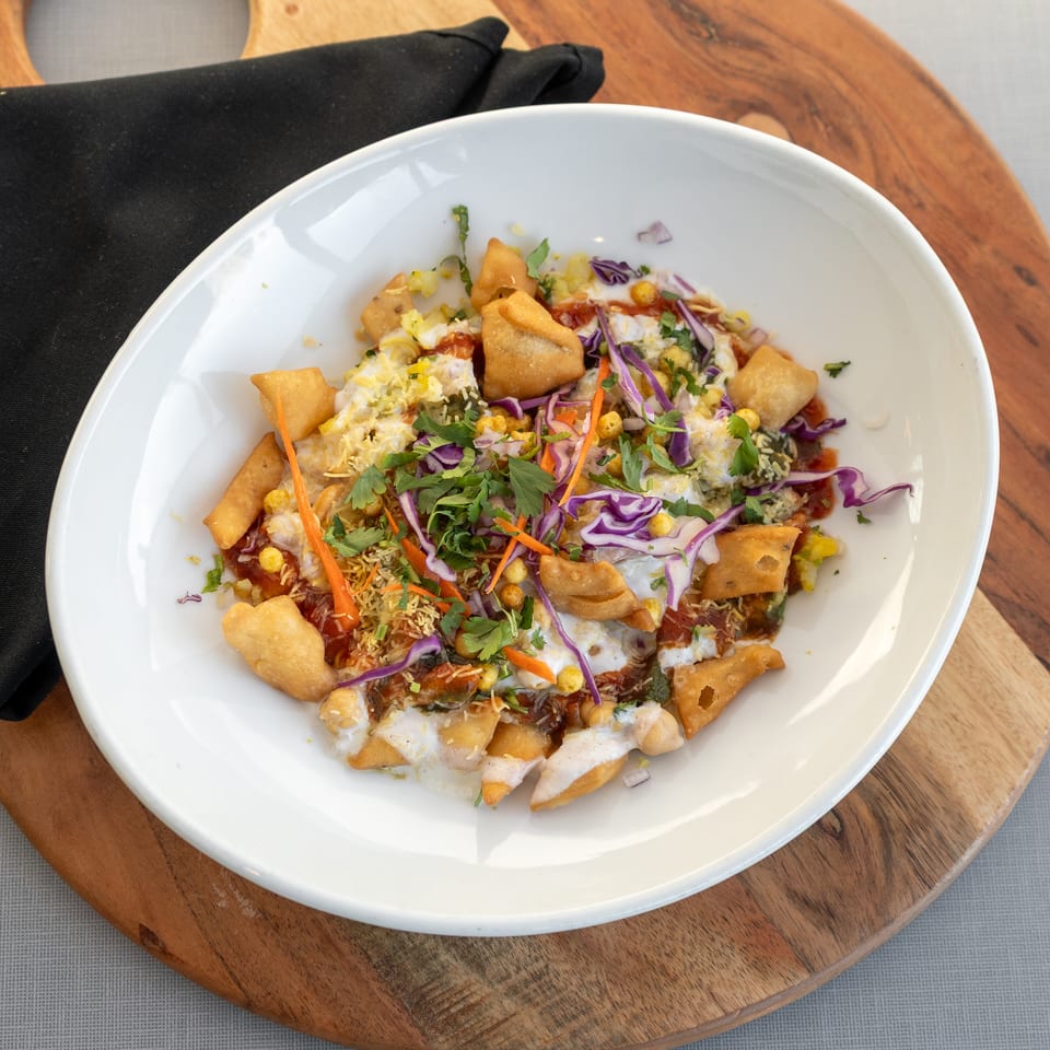 Indian chaat topped with crispy pastry, yogurt, chutney, sev, shredded cabbage, carrots, and fresh cilantro in a white bowl.