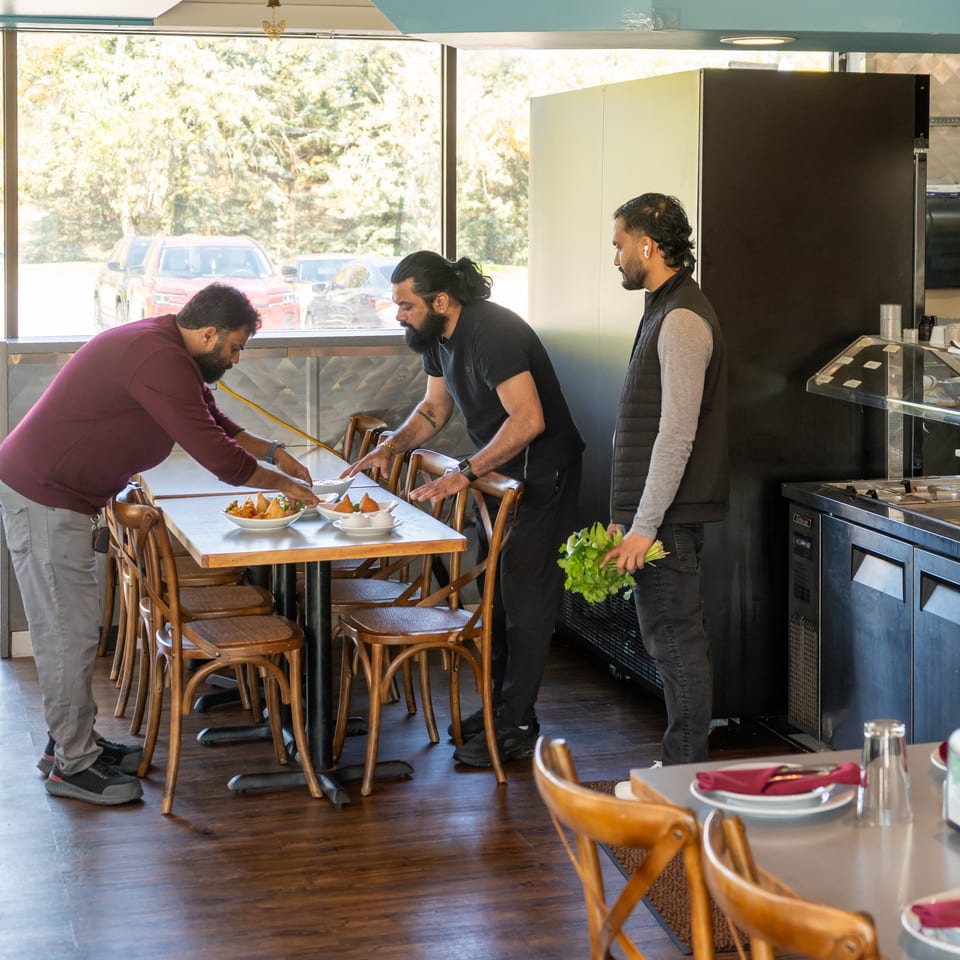 Three restaurant staff members arrange tables beside a display counter, with plated dishes and a bouquet of fresh herbs.