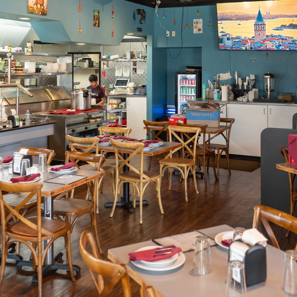 Restaurant dining room with turquoise walls, set tables, wooden chairs, open kitchen, and a television displaying Istanbul’s Galata Tower.