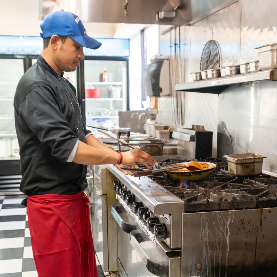 Restaurant cook in a blue cap and red apron prepares food in a commercial kitchen over a gas stove.